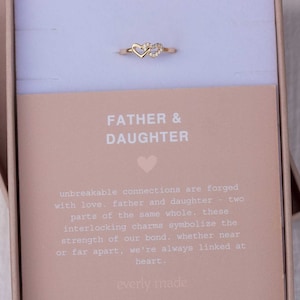 May include: A gold ring with two interlocking hearts, one with a small diamond accent. The ring is in a white box with a pink lid. The box has a card with the text "Father & Daughter" and a message about unbreakable connections.