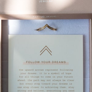 May include: A gold-colored ring with a double chevron design, presented in a jewelry box. The box contains a card with the text "FOLLOW YOUR DREAMS" and the brand name "everly made". The ring is a delicate piece of jewelry.