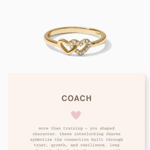 May include: A gold ring with two intertwined heart shapes, one set with small, clear gemstones. The ring is shown with a card bearing the word "COACH" and a message about trust and growth.