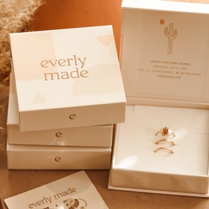 Pu&ograve; includere: Un set di tre anelli dorati in una scatola bianca con il nome del marchio "everly made" stampato sulla scatola. Gli anelli sono impilati uno sopra l'altro e sono realizzati in metallo sottile. La scatola &egrave; appoggiata su una superficie di legno.