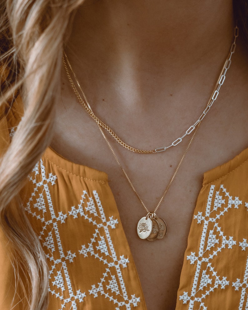 May include: A gold chain necklace with a delicate silver chain layered on top. The gold chain has three small, round pendants with a floral design.