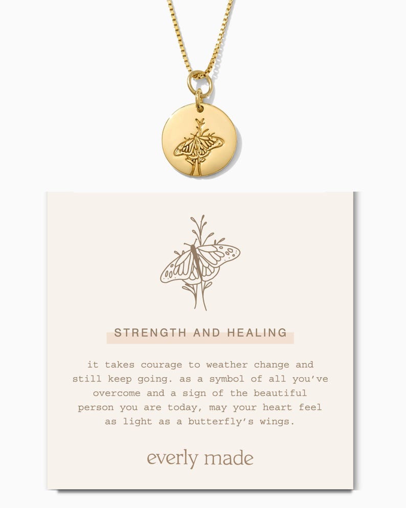 May include: Gold necklace with a round pendant featuring a butterfly design. The pendant is engraved with the words "Strength and Healing". The card below the necklace reads "it takes courage to weather change and still keep going. as a symbol of all you've overcome and a sign of the beautiful person you are today, may your heart feel as light as a butterfly's wings. everly made"