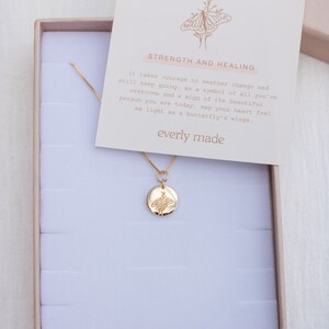 May include: A gold necklace with a delicate chain and a round pendant featuring a detailed butterfly design. The necklace comes in a small white box with a card that reads "Strength and Healing" and "everly made".