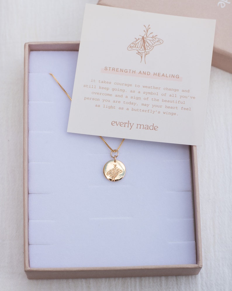 May include: A gold necklace with a delicate chain and a round pendant featuring a detailed butterfly design. The necklace comes in a small white box with a card that reads "Strength and Healing" and "everly made".