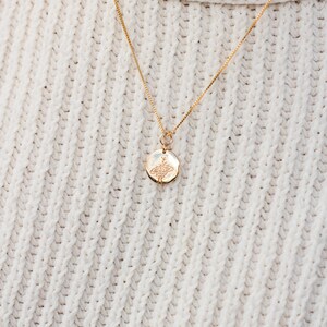 May include: A gold chain necklace with a small round pendant featuring a delicate floral design. The necklace is worn on a white knitted sweater.