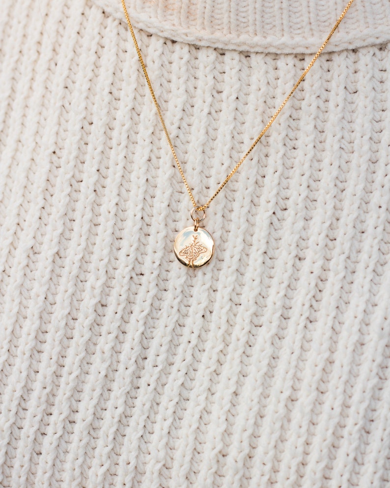 May include: A gold chain necklace with a small round pendant featuring a delicate floral design. The necklace is worn on a white knitted sweater.