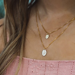 May include: Two gold chain necklaces with oval pendants. The top pendant is plain, while the bottom pendant features a delicate floral design.