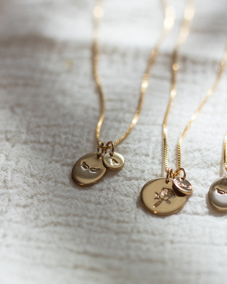 May include: Three gold necklaces with different charms. The first necklace has a small oval charm with a pair of wings, the second necklace has a larger oval charm with a rose design, and the third necklace has a small oval charm with a pair of wings.