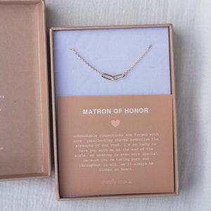 May include: A gold necklace with two interlocking charms in a gift box. The box has a card that reads "Matron of Honor" and a message about unbreakable connections.