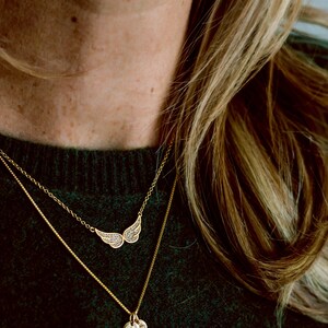 May include: A gold chain necklace with a small, round pendant and a larger pendant shaped like a pair of wings with small, sparkling stones. The necklace is worn over a dark green sweater.