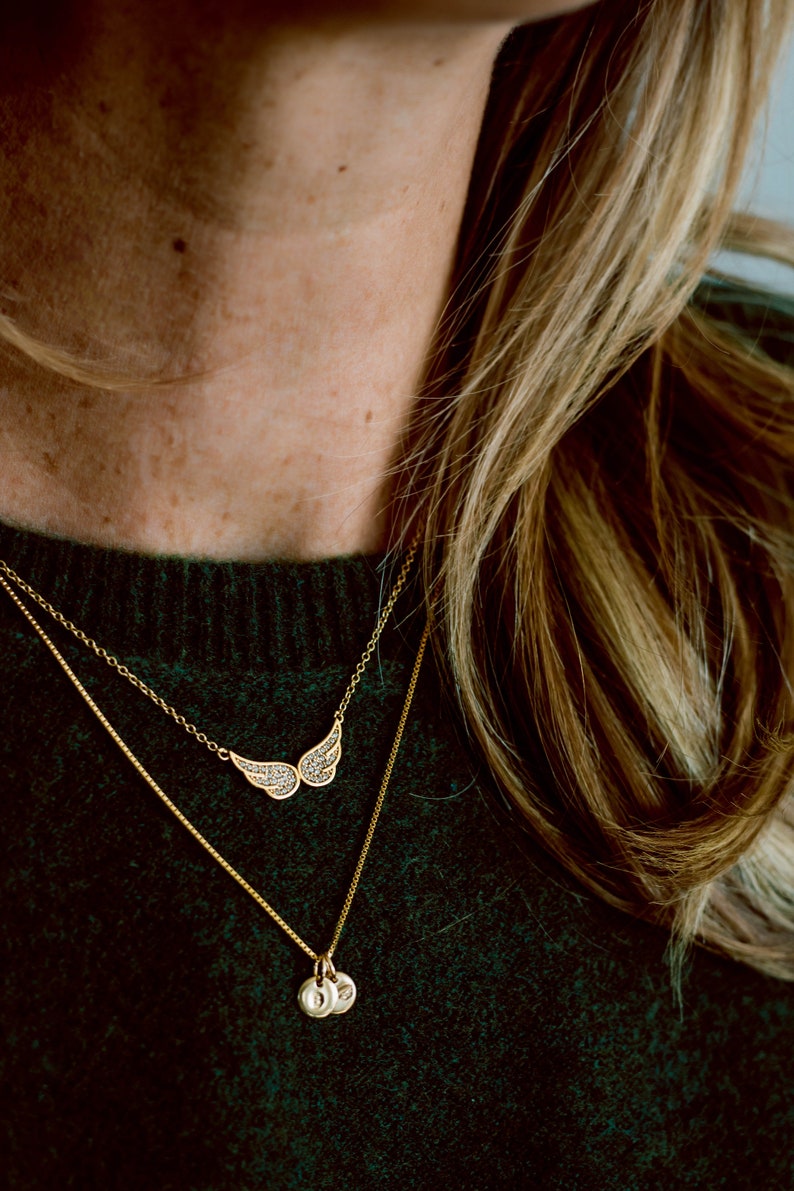 May include: A gold chain necklace with a small, round pendant and a larger pendant shaped like a pair of wings with small, sparkling stones. The necklace is worn over a dark green sweater.