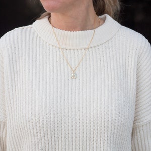 May include: A white cable knit sweater with a gold chain necklace and a small round pendant.