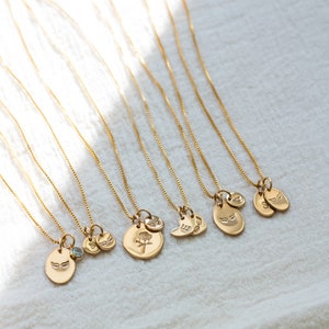 May include: A set of nine gold necklaces with different charms, including a heart, a flower, and wings. The necklaces are on a white fabric background.