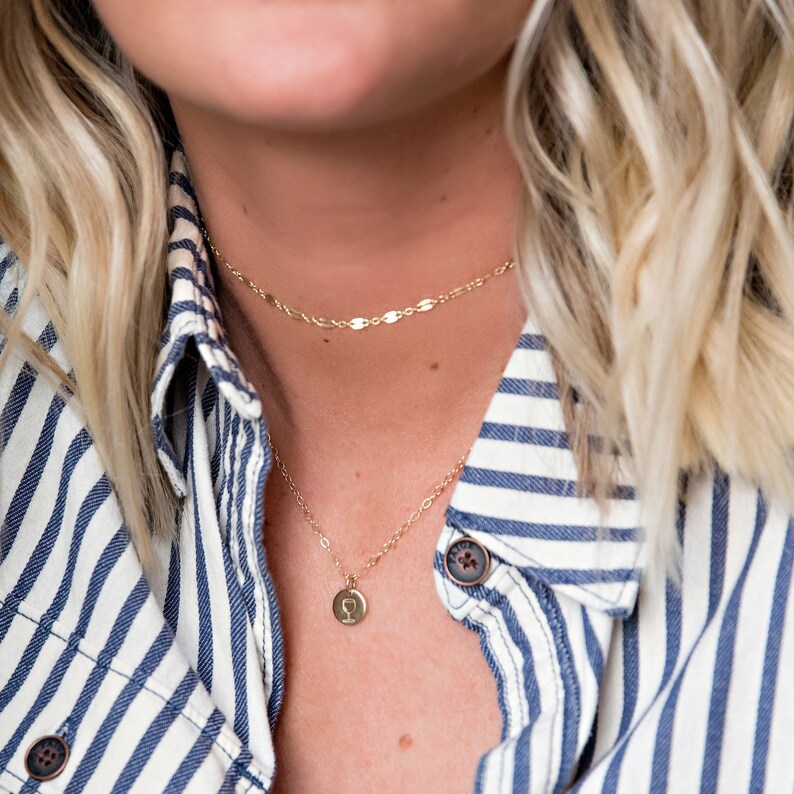 Pu&ograve; includere: Una collana a catena d'oro con un piccolo ciondolo d'oro con un design a forma di bicchiere di vino. La collana &egrave; indossata da una persona con i capelli biondi e una camicia a righe blu e bianche.
