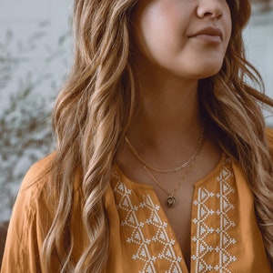 May include: A woman with long brown hair wearing a mustard yellow blouse with white embroidered geometric patterns. She is wearing a gold chain necklace with a small pendant.