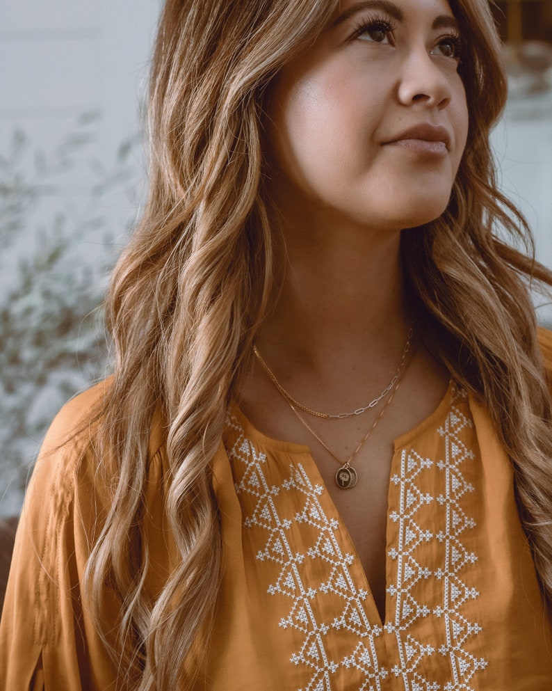 May include: A woman with long brown hair wearing a mustard yellow blouse with white embroidered geometric patterns. She is wearing a gold chain necklace with a small pendant.