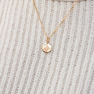 May include: A gold chain necklace with a small round pendant featuring a delicate floral design. The necklace is worn on a white knitted sweater.
