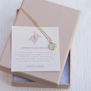 May include: A gold necklace with a delicate chain and a round pendant featuring a butterfly design. The pendant is on a light blue background. The necklace comes in a box with a card that reads "Strength and Healing" and includes a message about overcoming challenges and feeling light as a butterfly's wings. The card is signed "everly made".