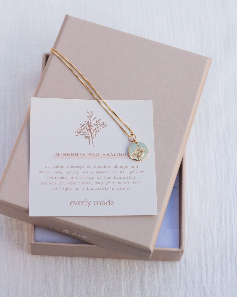 May include: A gold necklace with a delicate chain and a round pendant featuring a butterfly design. The pendant is on a light blue background. The necklace comes in a box with a card that reads "Strength and Healing" and includes a message about overcoming challenges and feeling light as a butterfly's wings. The card is signed "everly made".