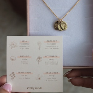 May include: A card with a floral design and the months of the year listed with corresponding flowers and their meanings. The card is next to a gold necklace with three charms, each featuring a different flower.