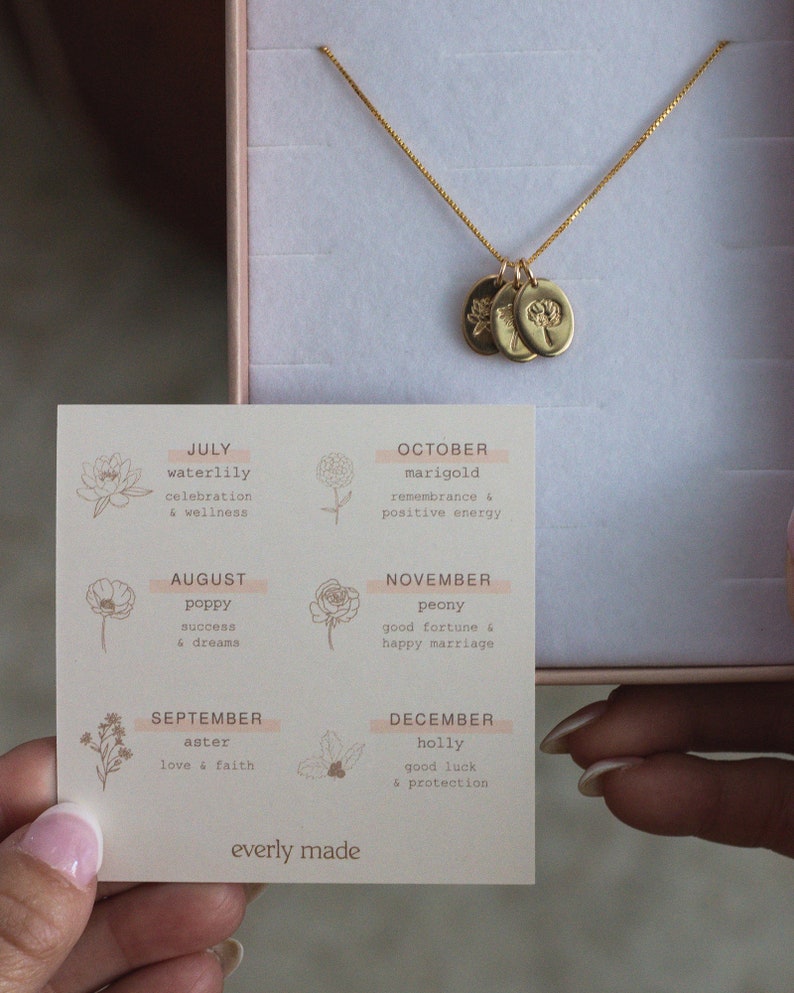 May include: A card with a floral design and the months of the year listed with corresponding flowers and their meanings. The card is next to a gold necklace with three charms, each featuring a different flower.
