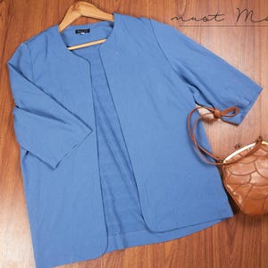 May include: A pale blue open-front jacket with three-quarter sleeves hangs on a wooden hanger. A brown leather purse with a distinctive, textured design sits nearby. The jacket and purse are on a wooden surface.