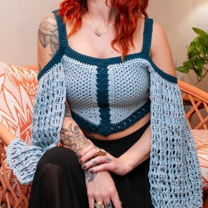 Pode incluir: Um top de crochê azul e turquesa com um design de ombro caído. O top é curto e tem um caimento solto. A modelo está usando o top com uma saia preta.
