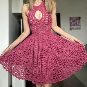 May include: A magenta-colored, halter-style crochet dress. The dress features a keyhole detail at the bust and a flared skirt. The open weave design is visible throughout the garment, creating a textured appearance. The dress is a summer or beachwear item.