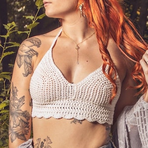 May include: A woman with bright red hair wearing a white crocheted halter top, blue jeans with embroidery, and a white crocheted shawl. She has tattoos on her arms and a gold necklace.
