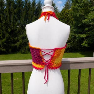 May include: A multi-coloured crochet halter top featuring a vibrant gradient of yellow, orange, red, and magenta yarn. The garment includes a lace-up back closure with matching yarn ties and a halter neck strap that secures at the back of the neck.