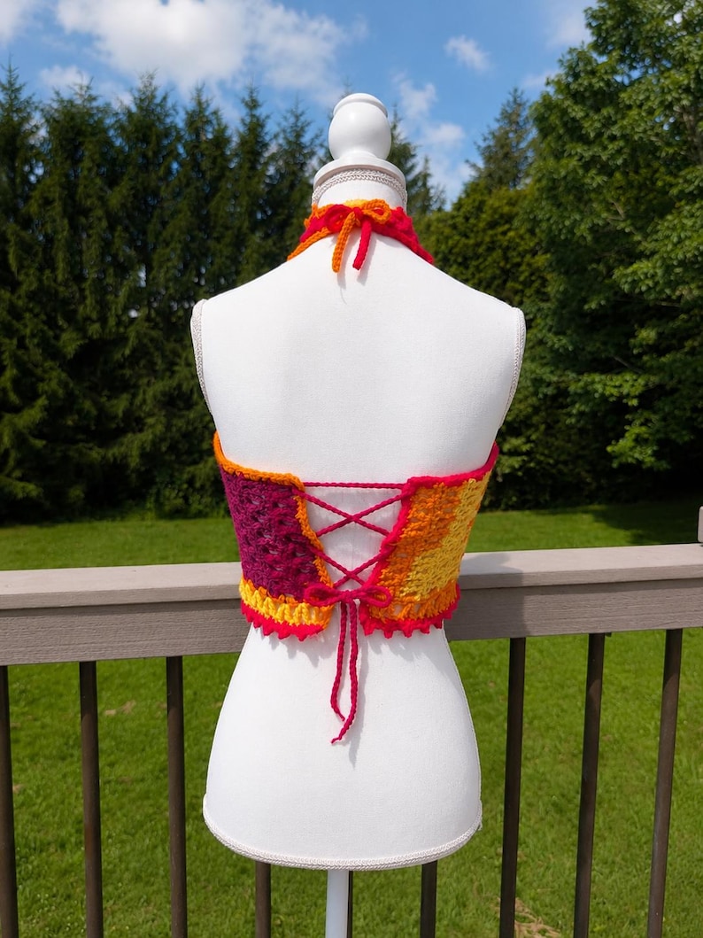 May include: A multi-coloured crochet halter top featuring a vibrant gradient of yellow, orange, red, and magenta yarn. The garment includes a lace-up back closure with matching yarn ties and a halter neck strap that secures at the back of the neck.