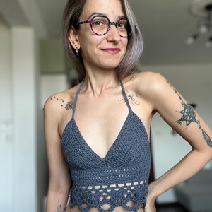 May include: A gray crochet halter top with a V-neck and scalloped bottom edge. The top is handmade and appears to be a summer garment. The top is paired with blue jeans.