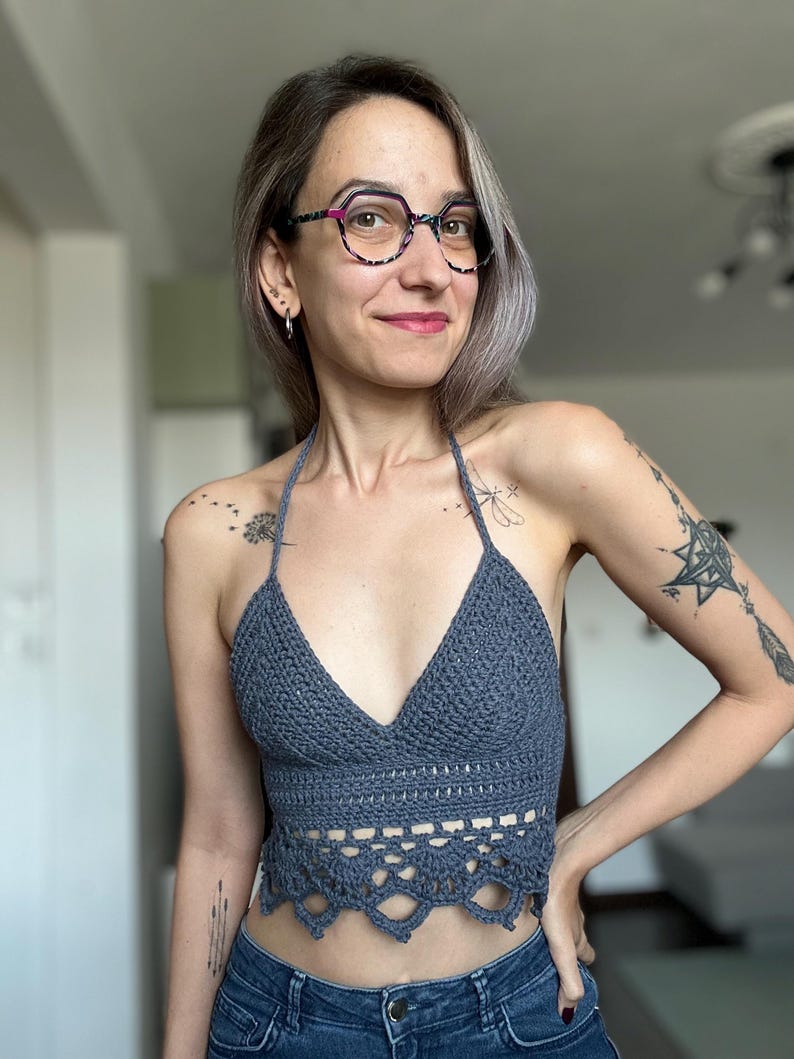 May include: A gray crochet halter top with a V-neck and scalloped bottom edge. The top is handmade and appears to be a summer garment. The top is paired with blue jeans.