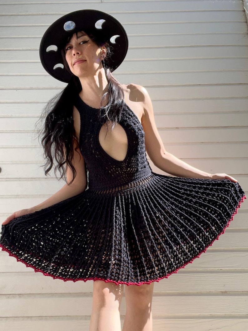 May include: A black crochet dress with a halter neckline and a flared skirt. The dress has a keyhole cutout at the chest and a red trim along the hem. A black hat with white crescent moon accents completes the look.