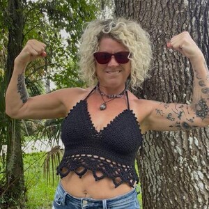 May include: A black crochet halter top with a scalloped edge. The top is paired with blue denim shorts. The image is taken outdoors with a tree trunk in the background.
