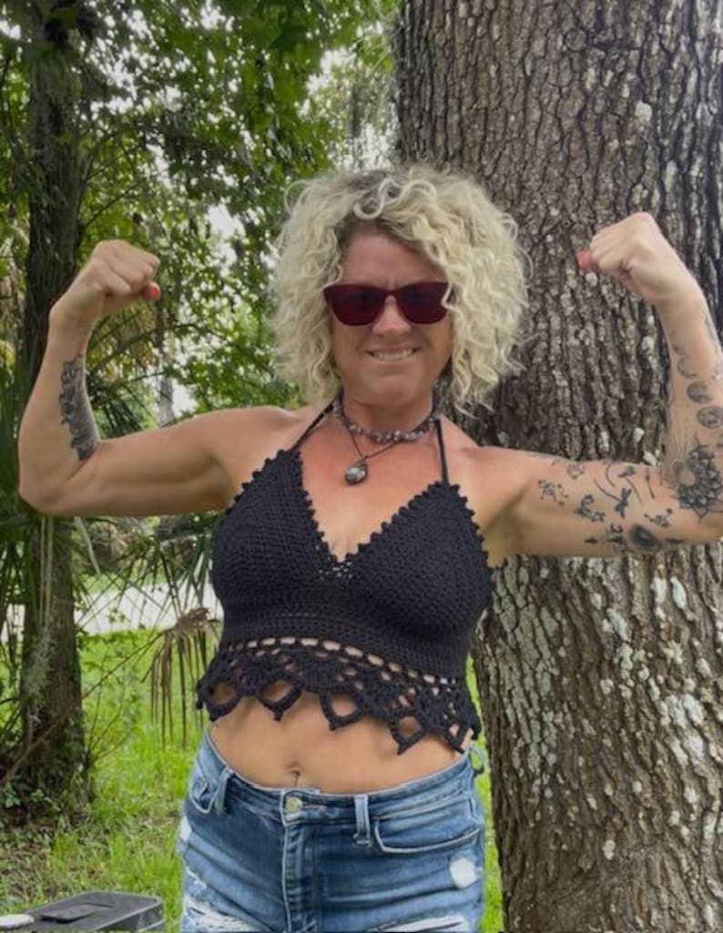 May include: A black crochet halter top with a scalloped edge. The top is paired with blue denim shorts. The image is taken outdoors with a tree trunk in the background.