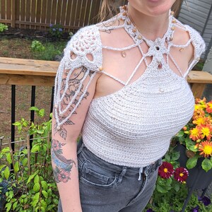 May include: A white crochet top with a beaded neckline and crocheted shoulder straps. The top has a geometric design and is worn with dark gray jeans.