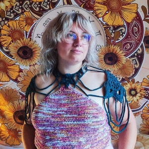 May include: A woman wearing a multi-colored crochet halter top with matching crocheted shoulder straps. The top has a tie closure at the back of the neck. The woman is standing in front of a tapestry with a sunflower pattern.
