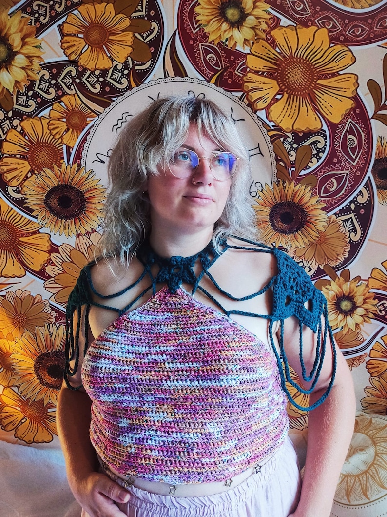 May include: A woman wearing a multi-colored crochet halter top with matching crocheted shoulder straps. The top has a tie closure at the back of the neck. The woman is standing in front of a tapestry with a sunflower pattern.