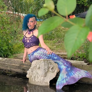May include: A purple crochet halter top with a scalloped hem and a matching purple and blue mermaid tail featuring a skeletal rib cage print. The tail has a long, flowing fin and a gradient colour pattern transitioning from deep purple to light blue.
