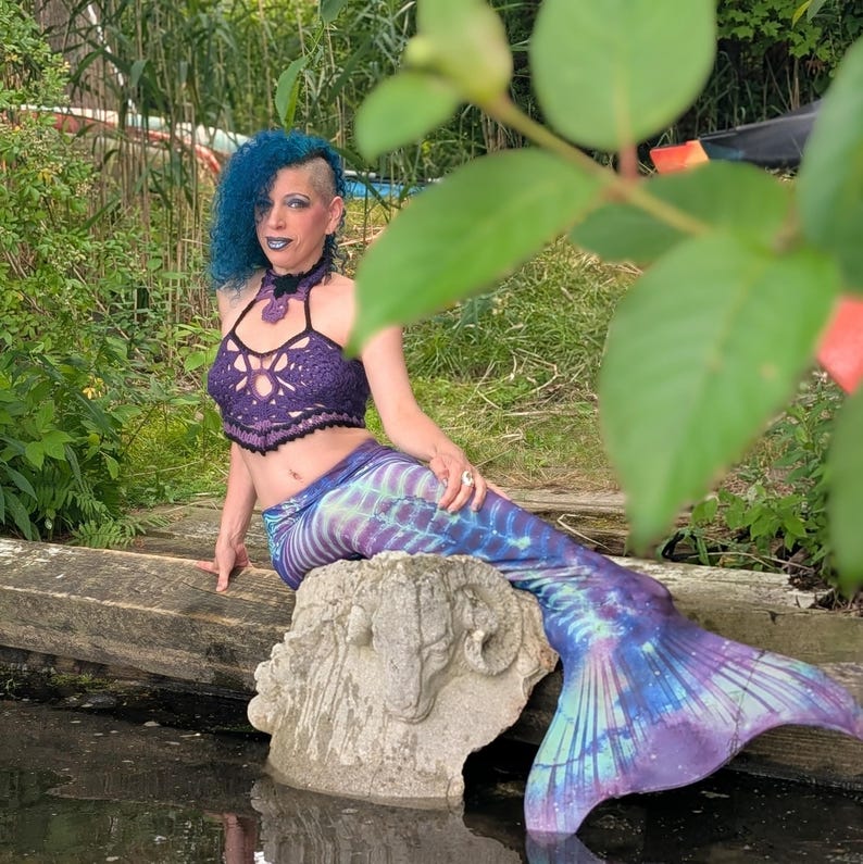 May include: A purple crochet halter top with a scalloped hem and a matching purple and blue mermaid tail featuring a skeletal rib cage print. The tail has a long, flowing fin and a gradient colour pattern transitioning from deep purple to light blue.
