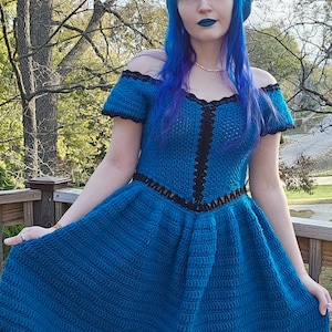 May include: A handmade, off-the-shoulder, blue crochet dress with black trim. The dress features a fitted bodice and a flared skirt. The model is wearing a matching blue beret and has blue hair and lipstick. The dress is a unique, handcrafted garment.