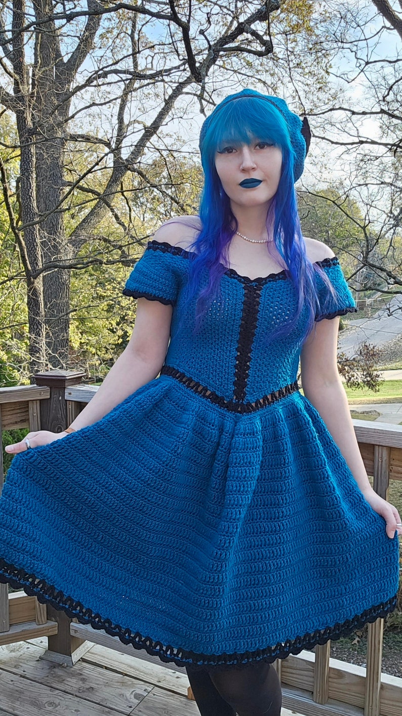 May include: A handmade, off-the-shoulder, blue crochet dress with black trim. The dress features a fitted bodice and a flared skirt. The model is wearing a matching blue beret and has blue hair and lipstick. The dress is a unique, handcrafted garment.