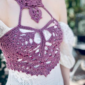 May include: A mauve crochet halter top featuring an intricate openwork pattern and a high neck collar. The garment is embellished with small, matching purple beads woven throughout the yarn, creating a textured design that sits over a white lace dress.