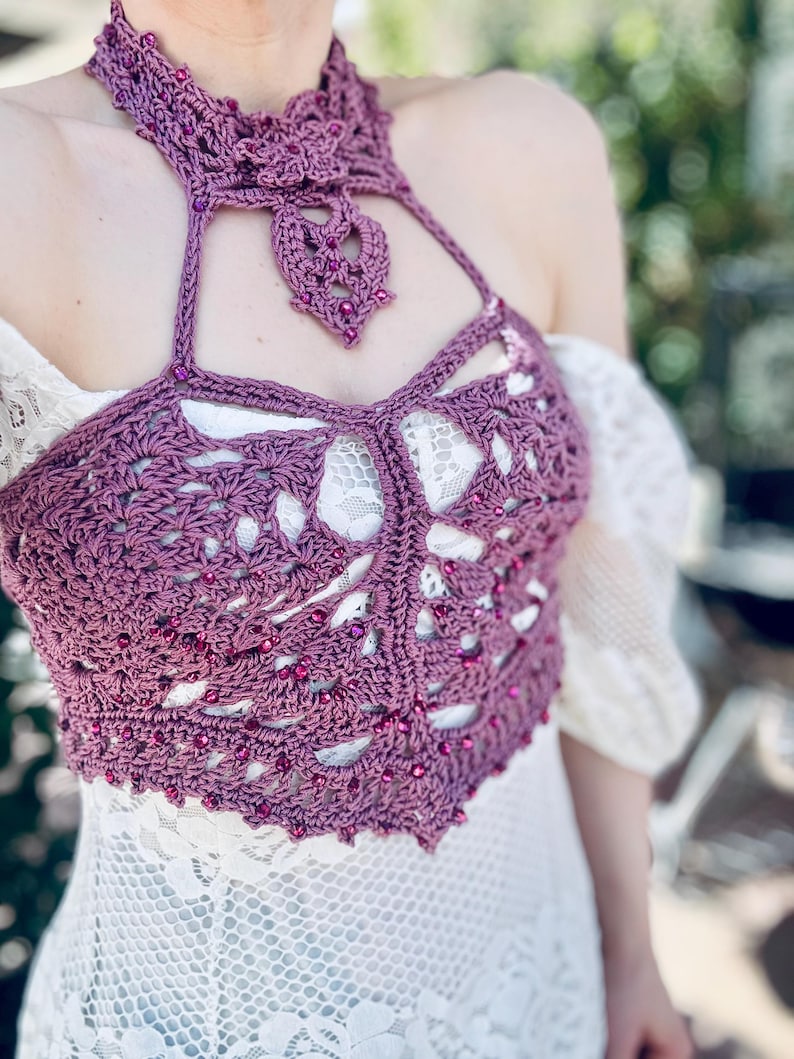 May include: A mauve crochet halter top featuring an intricate openwork pattern and a high neck collar. The garment is embellished with small, matching purple beads woven throughout the yarn, creating a textured design that sits over a white lace dress.