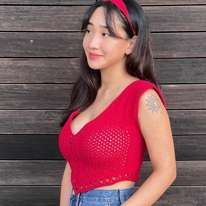 May include: A red crochet crop top with a deep V-neck and a matching red headband. The top is sleeveless and has a textured, open weave design. The model is wearing blue jeans.