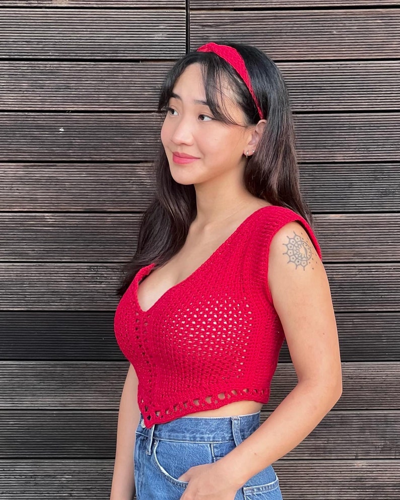 May include: A red crochet crop top with a deep V-neck and a matching red headband. The top is sleeveless and has a textured, open weave design. The model is wearing blue jeans.