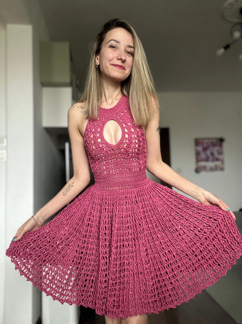 May include: A magenta-colored, halter-style crochet dress. The dress features a keyhole detail at the bust and a flared skirt. The open weave design is visible throughout the garment, creating a textured appearance. The dress is a summer or beachwear item.