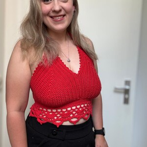 May include: A red halter top with a crocheted design. The top features a scalloped edge at the waist and a halter neck. The top is paired with black pants. The top is handmade.