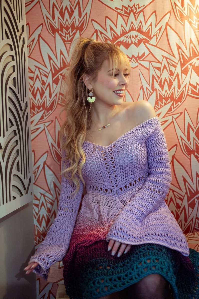 May include: A lavender crochet dress with long sleeves and a flared skirt, transitioning to shades of pink, red, and green. The off-the-shoulder neckline and intricate stitchwork create a bohemian aesthetic. The model is wearing gold earrings and a necklace.